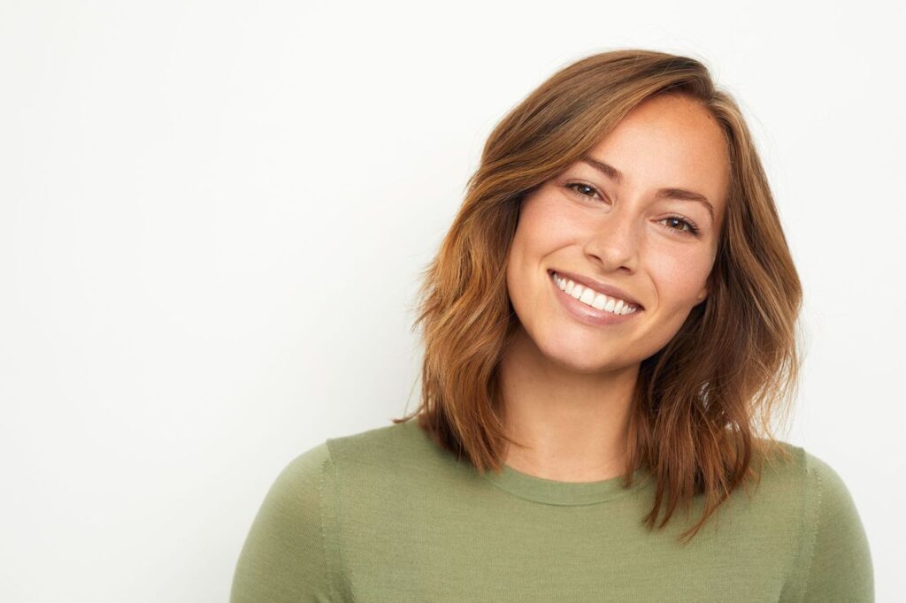 Young woman with brown hair, wearing an olive green sweater, smiles widely to show her new veneers